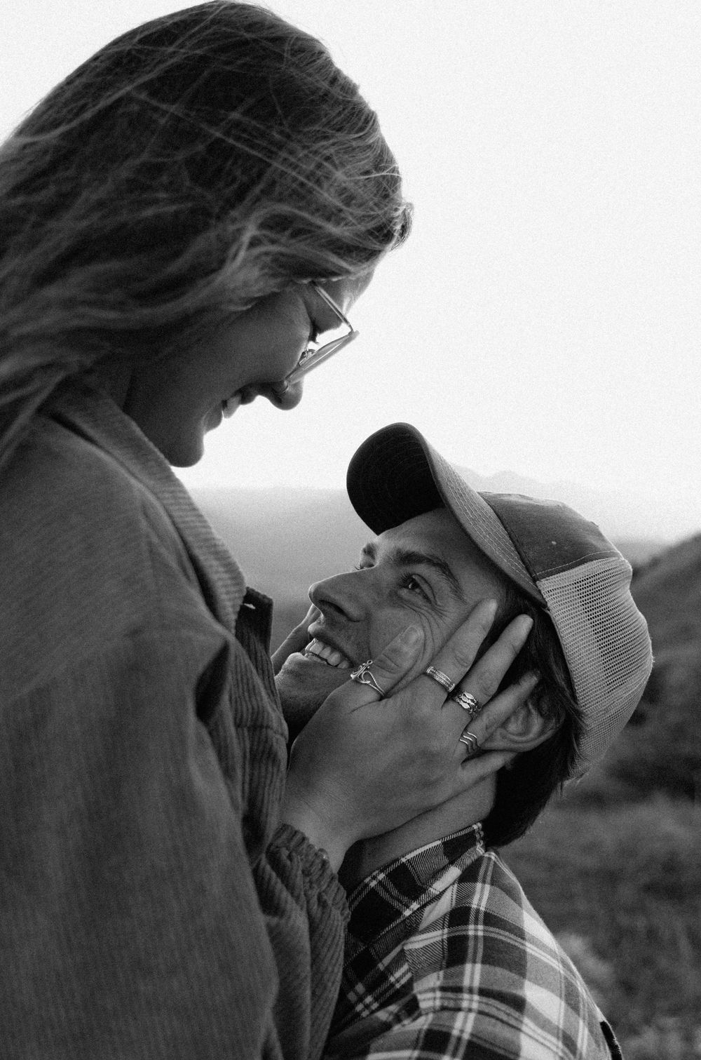 A black and white candid photo of a romantic moment between a couple outdoors, with one wearing a baseball cap.