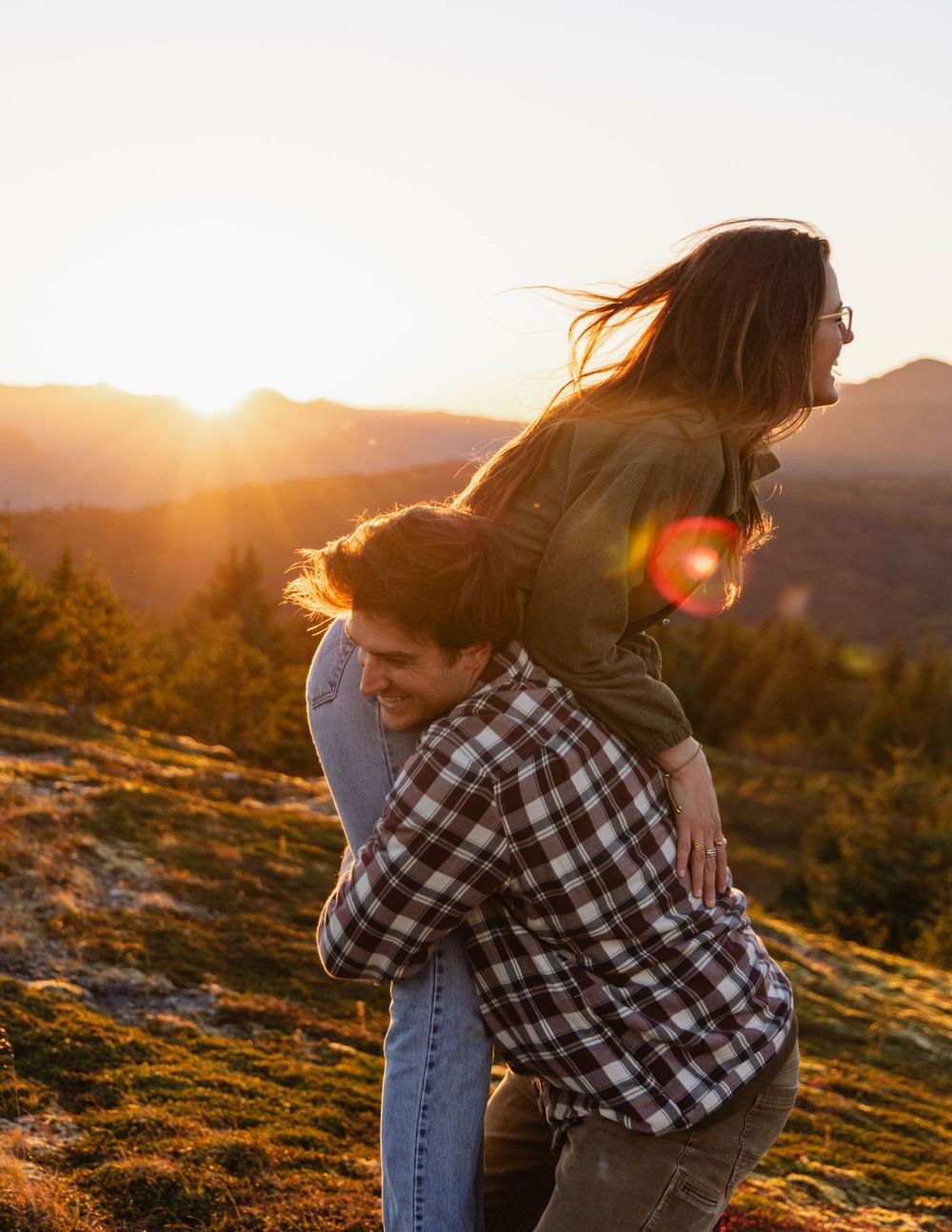 A couple shares a joyful moment at sunset on a mountainside, with sunlight streaming through long flowing hair.