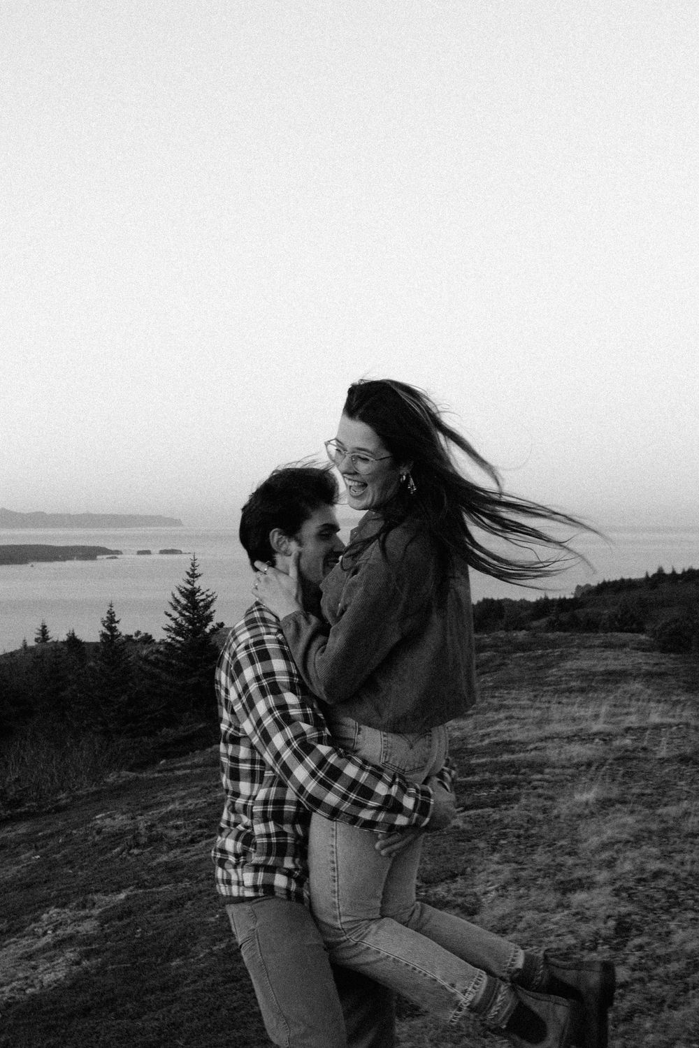 A couple embraces and kisses on a windswept coastal hillside in black and white.