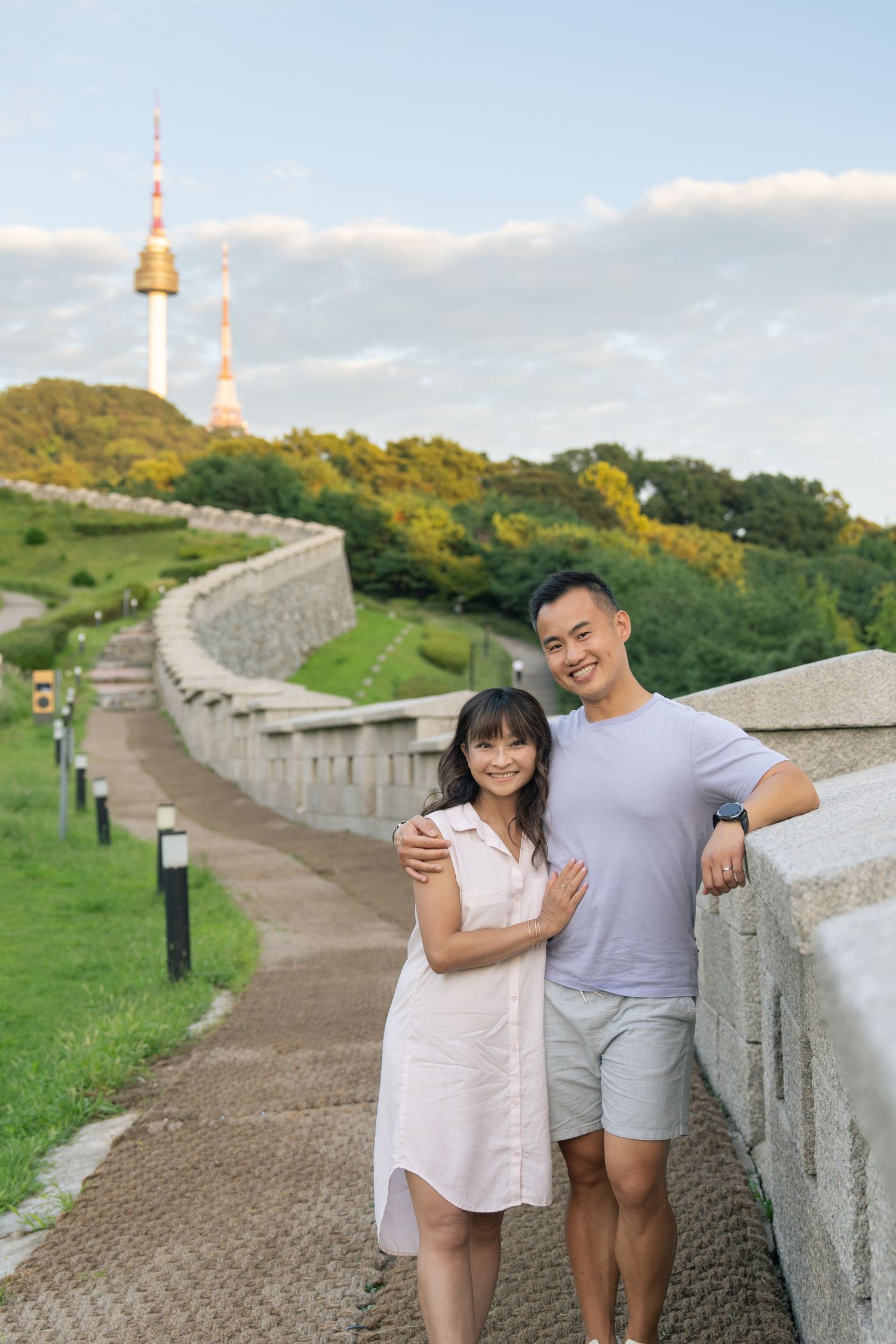 Couple standing along stone wall path with N Seoul Tower and lush greenery visible in background.