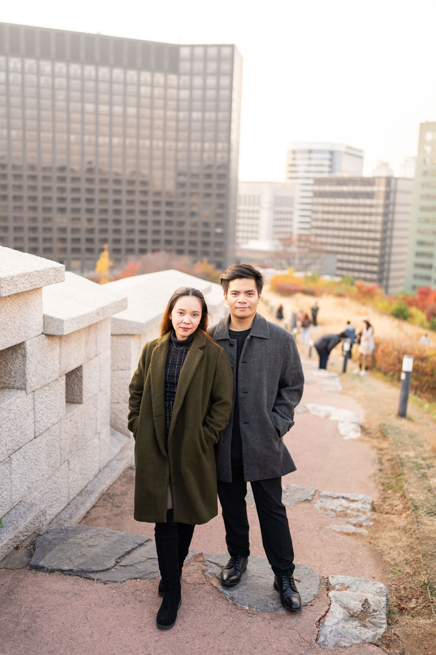 Two figures stand together on an urban path with tall office buildings visible in the background.