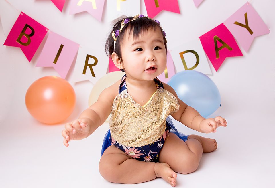 Baby wearing gold outfit during birthday photoshoot with pink decorations and balloons.