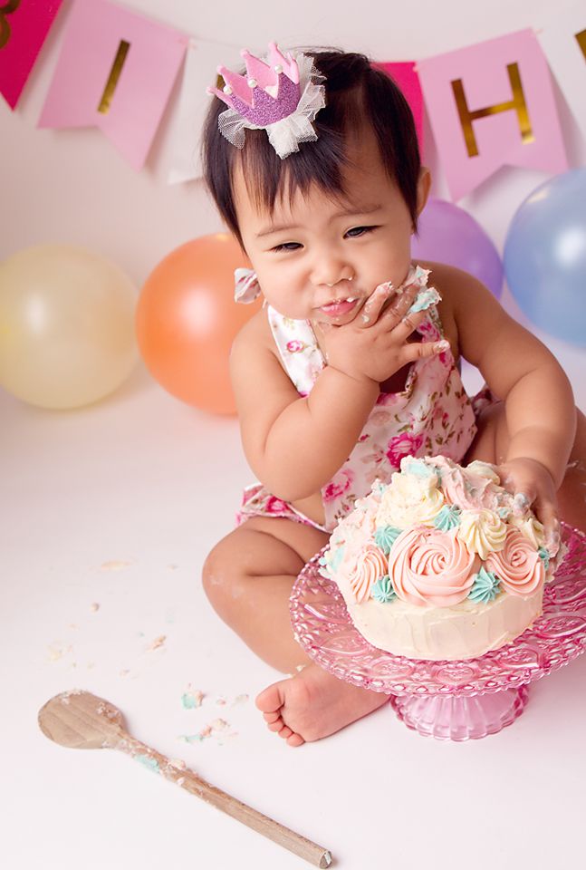 Baby in pink dress enjoying first birthday cake smash session with colorful balloons and decorations.