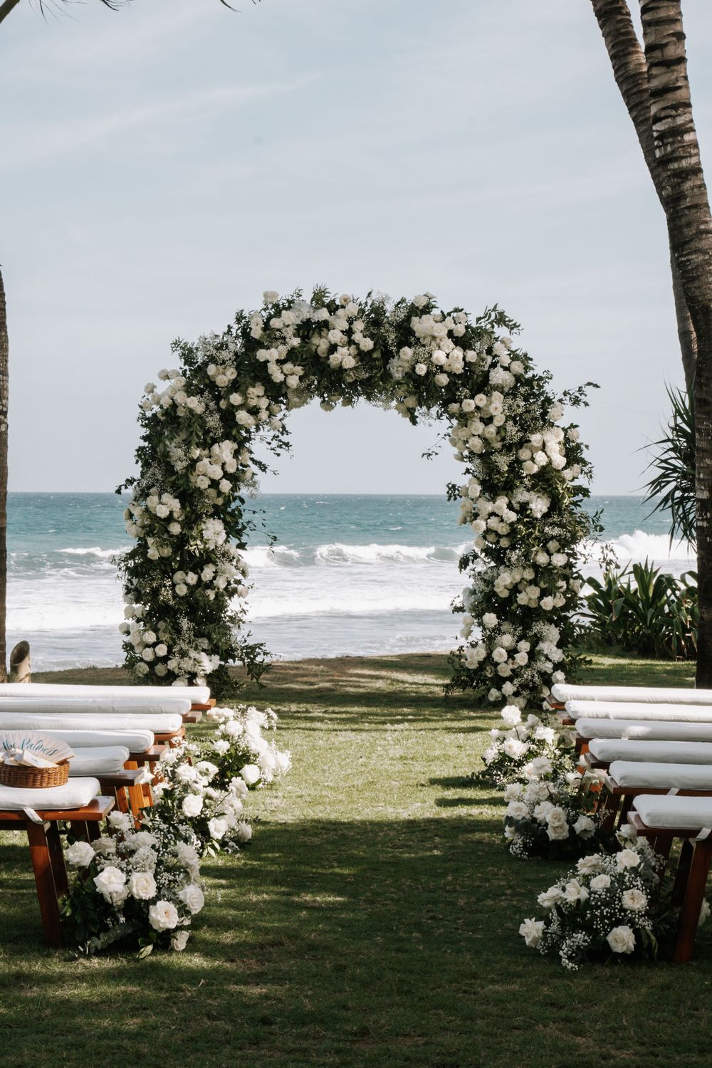 White floral arches line a beachfront wedding ceremony setup with wooden benches and ocean views.