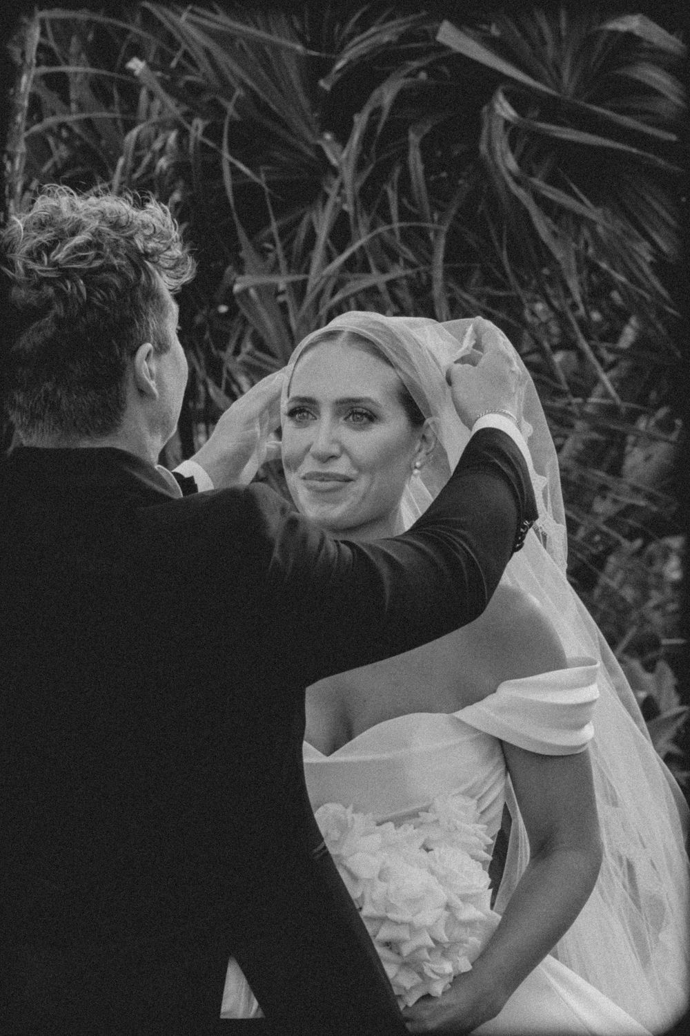 Black and white wedding photo showing tender moment between couple as veil is adjusted against foliage background.