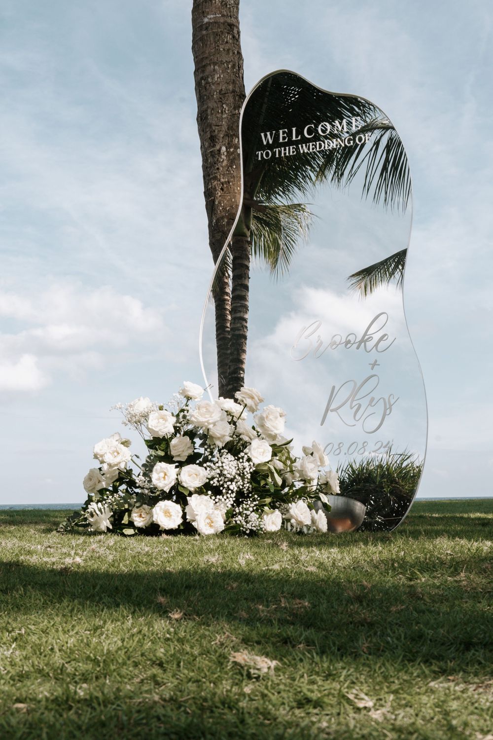 Modern wedding welcome sign with acrylic panels and white floral arrangements displayed on grass near palm trees.