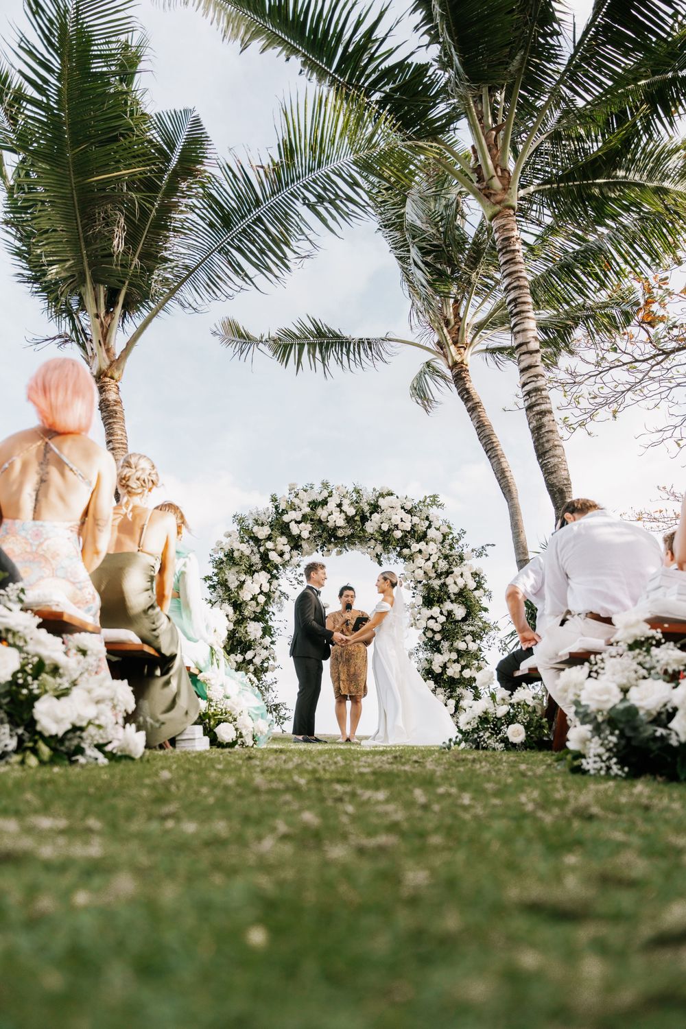 A wedding ceremony scene on a grassy hill with palm trees and white floral arrangements against a blue sky.