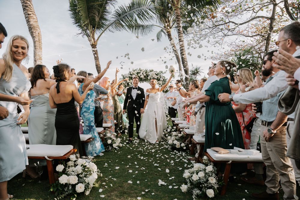 Wedding guests celebrate joyfully outdoors under palm trees with confetti flying through the air at a tropical celebration.