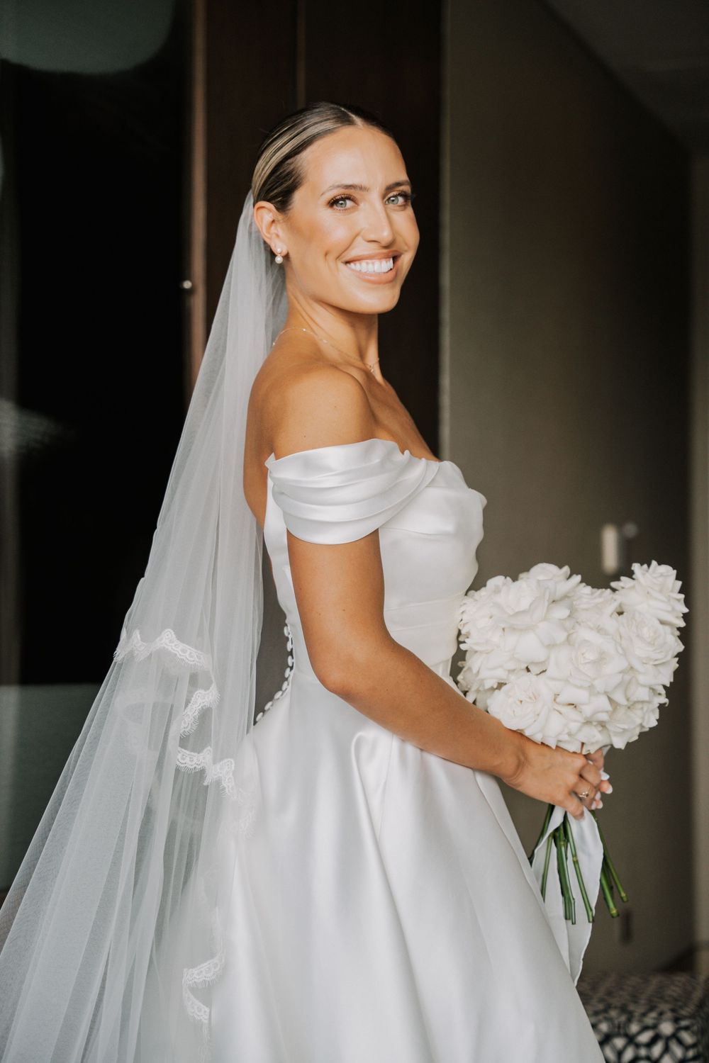 Elegant bride in classic white wedding gown holds white floral bouquet while wearing long flowing veil.