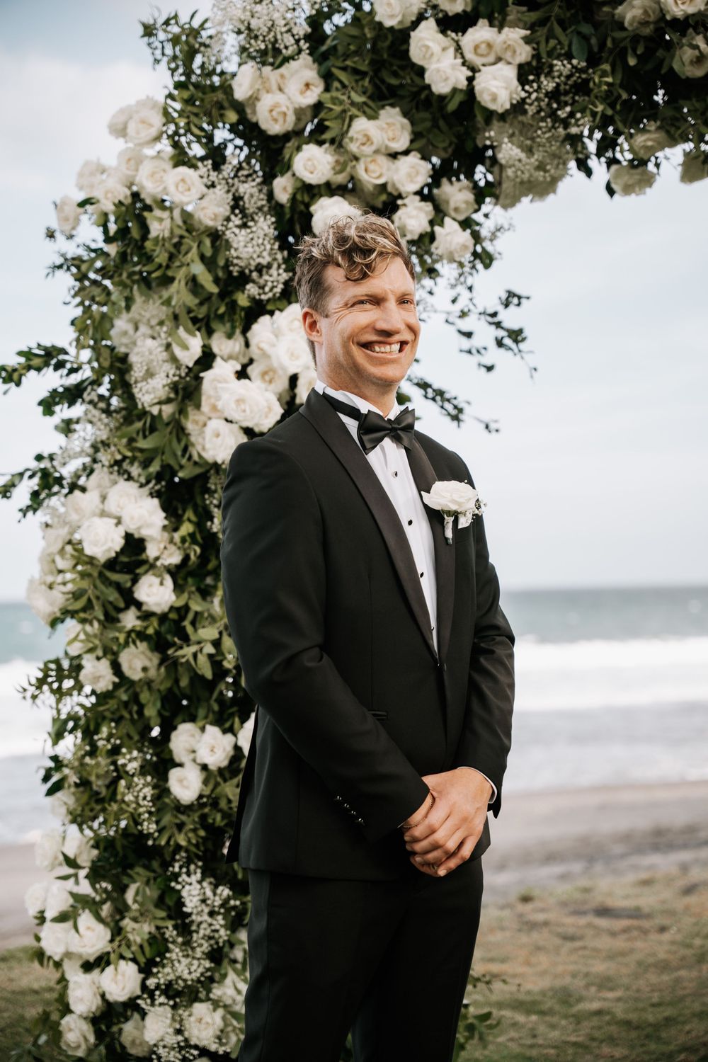 A groom in a black tuxedo smiles joyfully under a floral arch with white roses against a misty beach backdrop.