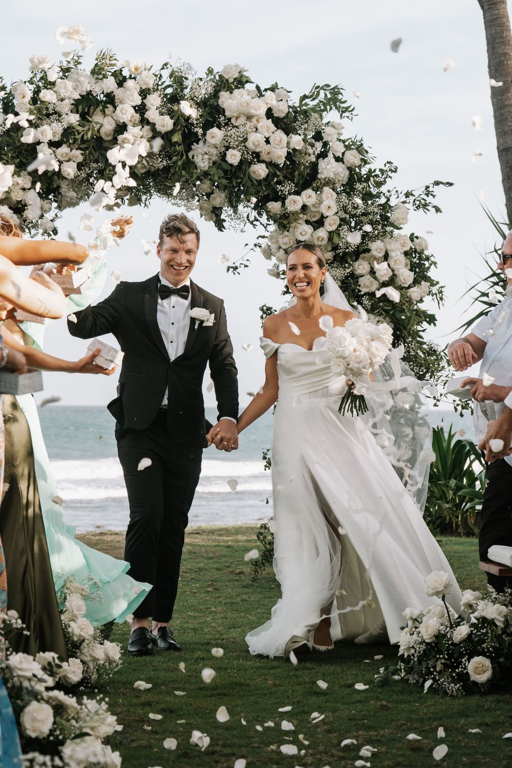 A joyful couple walks down the aisle under a stunning white floral arch at their oceanfront wedding ceremony.