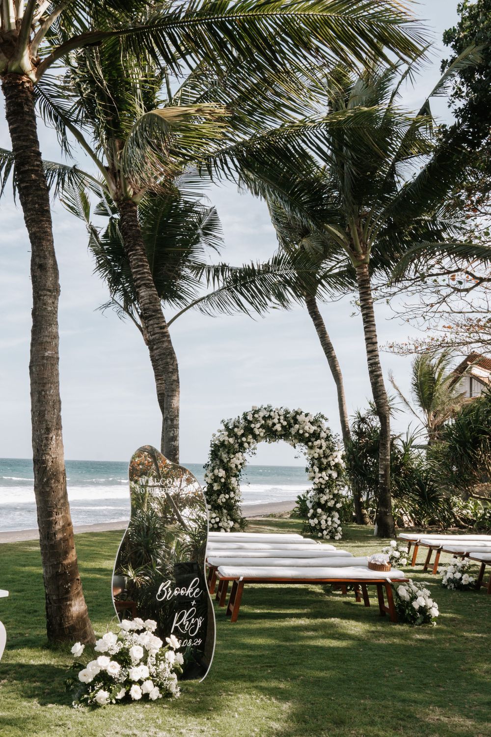 Tropical beach wedding ceremony setup with palm trees, white floral arrangements and ocean views in the background.
