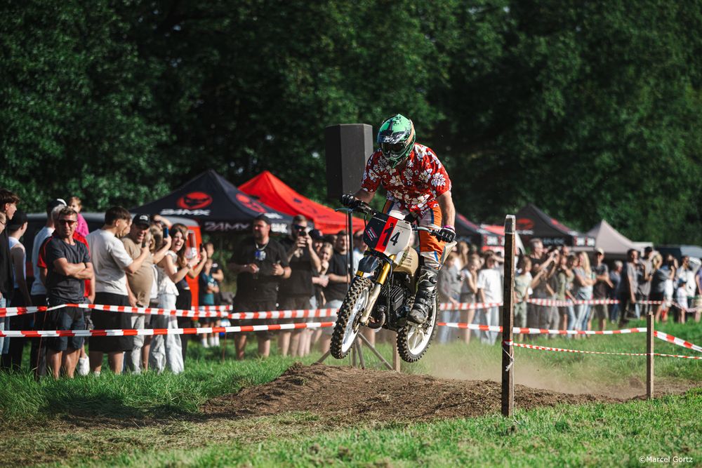 A motocross rider performs a jump on a dirt track while spectators watch from behind barriers at an outdoor event.