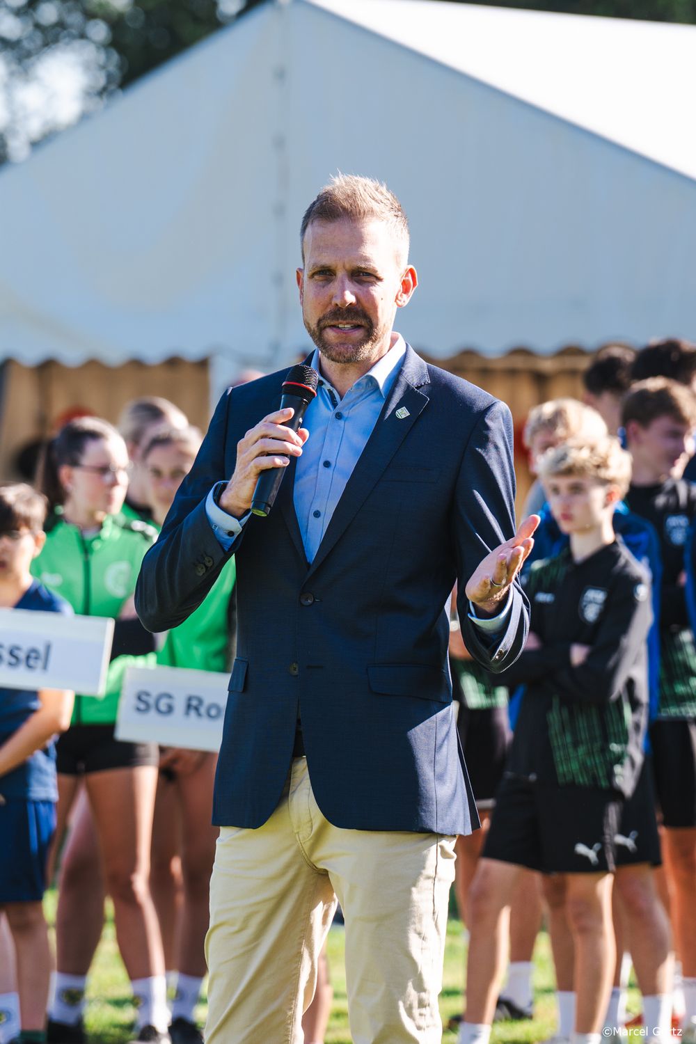 An official in a navy blazer and light pants speaks into a microphone at an outdoor sporting event.