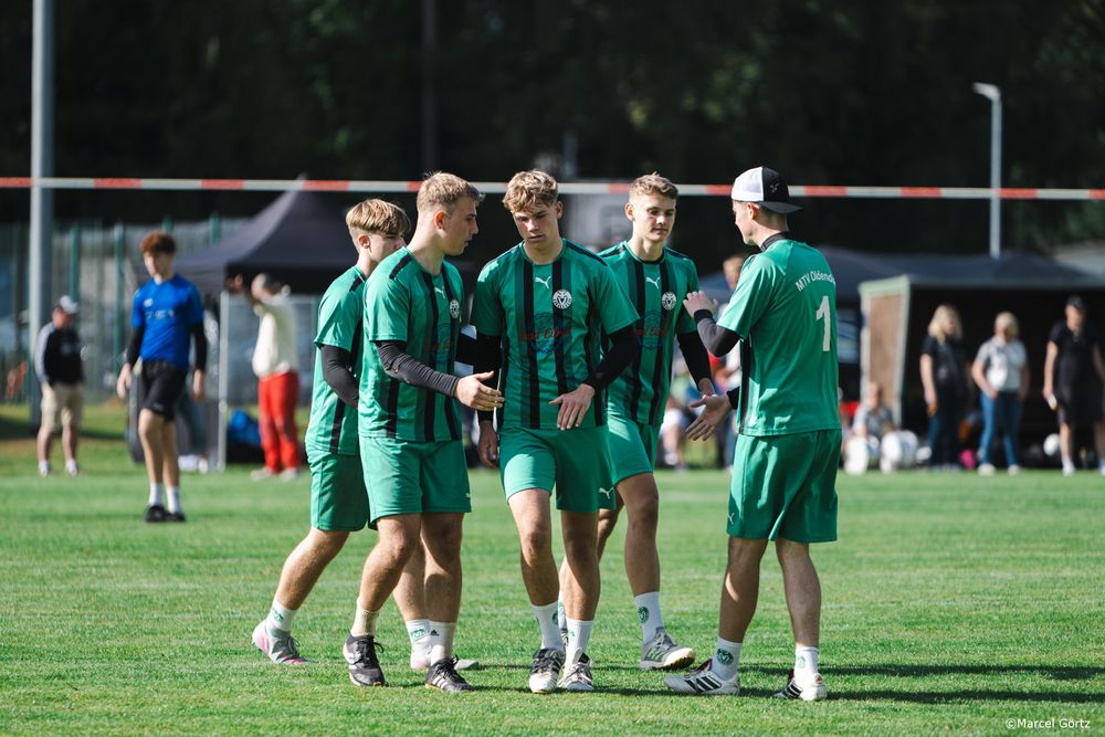 Soccer players in green uniforms gather on a grass field during practice or warm-up session.