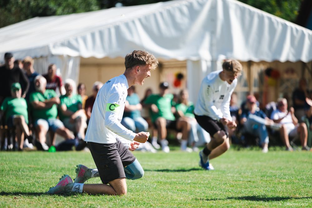 Two children in white shirts play on a grassy field with a white tent and spectators in the background.