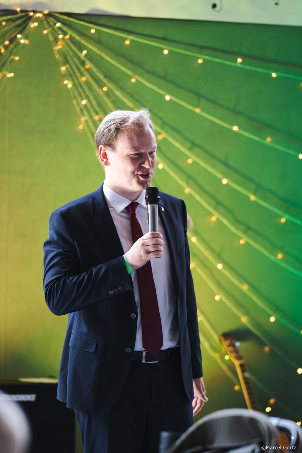 A speaker in a navy suit and red tie performs against a green backdrop with string lights.