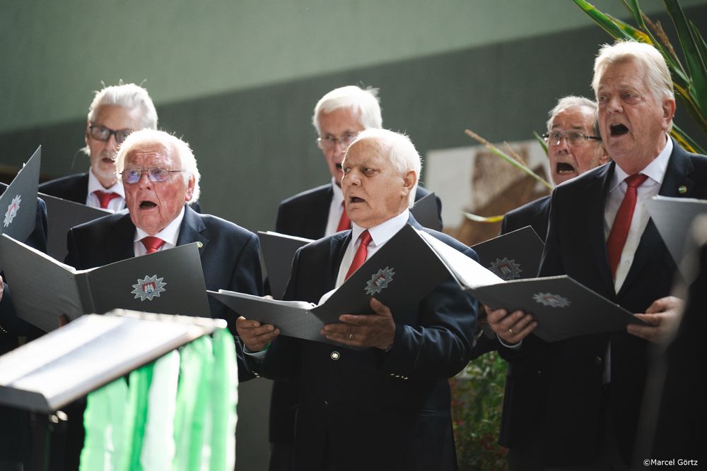 A group of men in dark suits singing together in a formal choir performance.