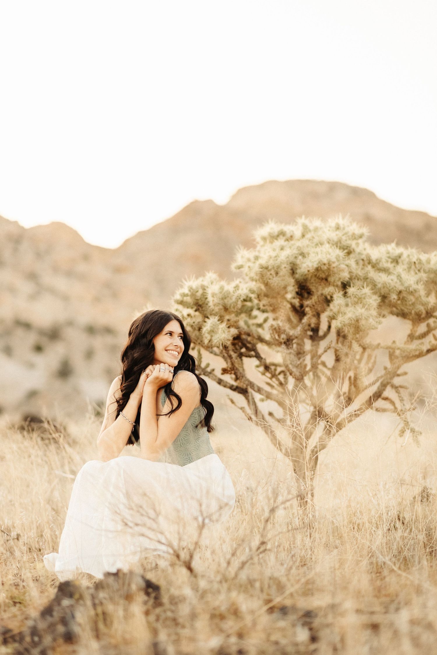 A person in a white dress reclines in a desert landscape with Joshua trees and golden light at sunset.