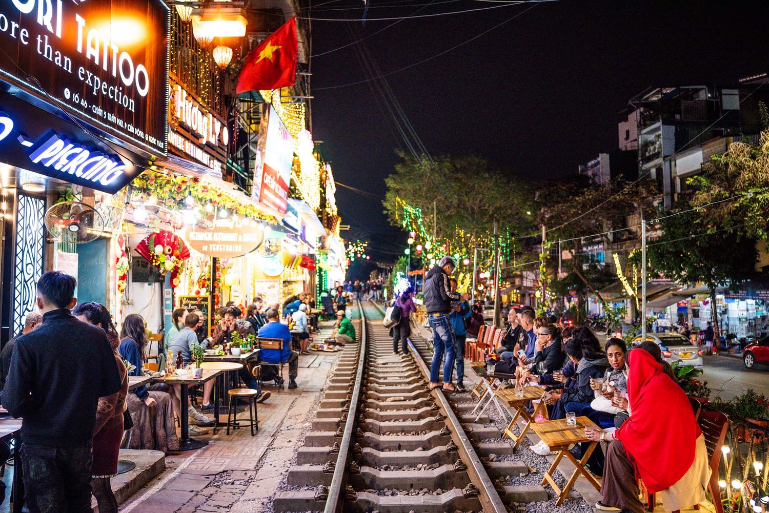 Vibrant nighttime scene of an outdoor restaurant patio with string lights illuminating diners along train tracks.