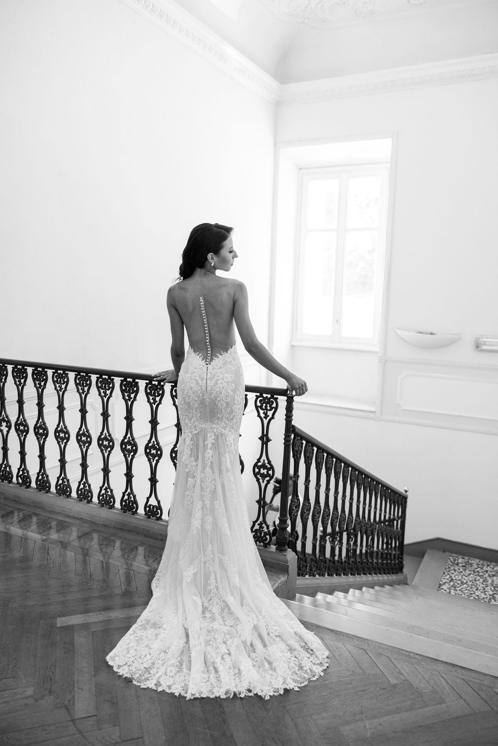 Elegant lace wedding dress with flowing train shown from behind in a bright interior space with ornate railing.