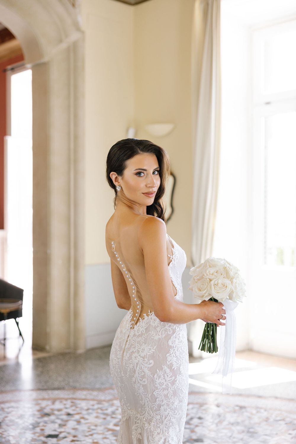Elegant lace wedding dress with intricate details and sheer back photographed in a bright indoor setting.