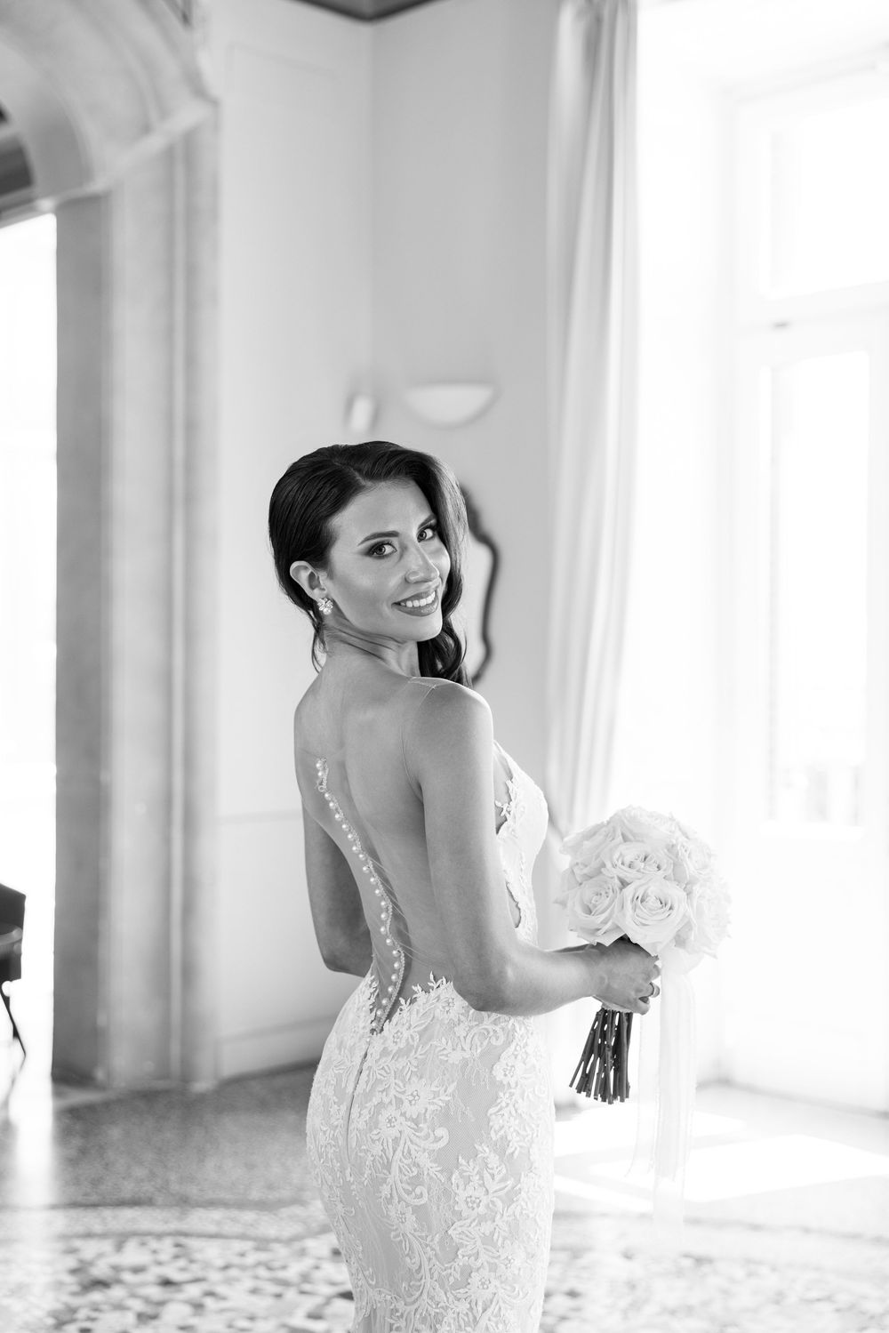 Bride wearing an elegant lace wedding dress poses by a window in black and white photography.