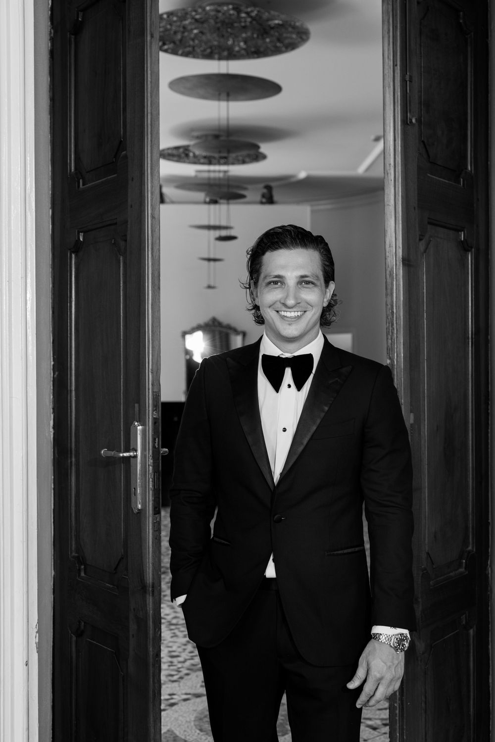 Groomsmen in black tuxedos pose in an elegant hallway with ornate ceiling fixtures.