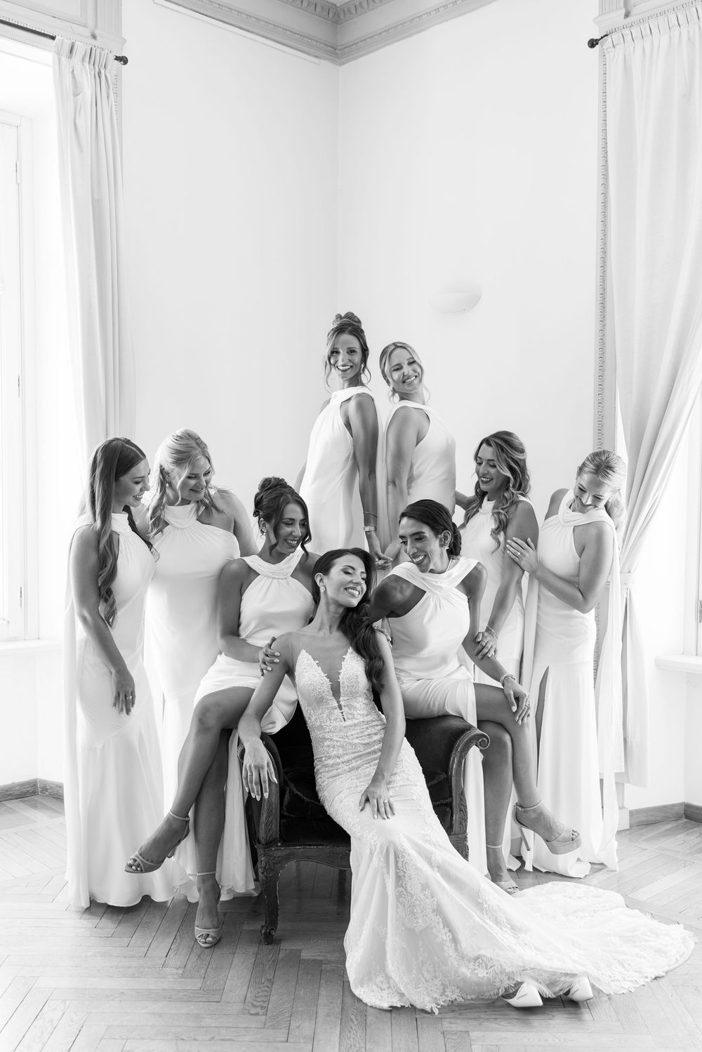 Bridesmaids in white dresses pose playfully in an elegant room with white curtains and natural light.