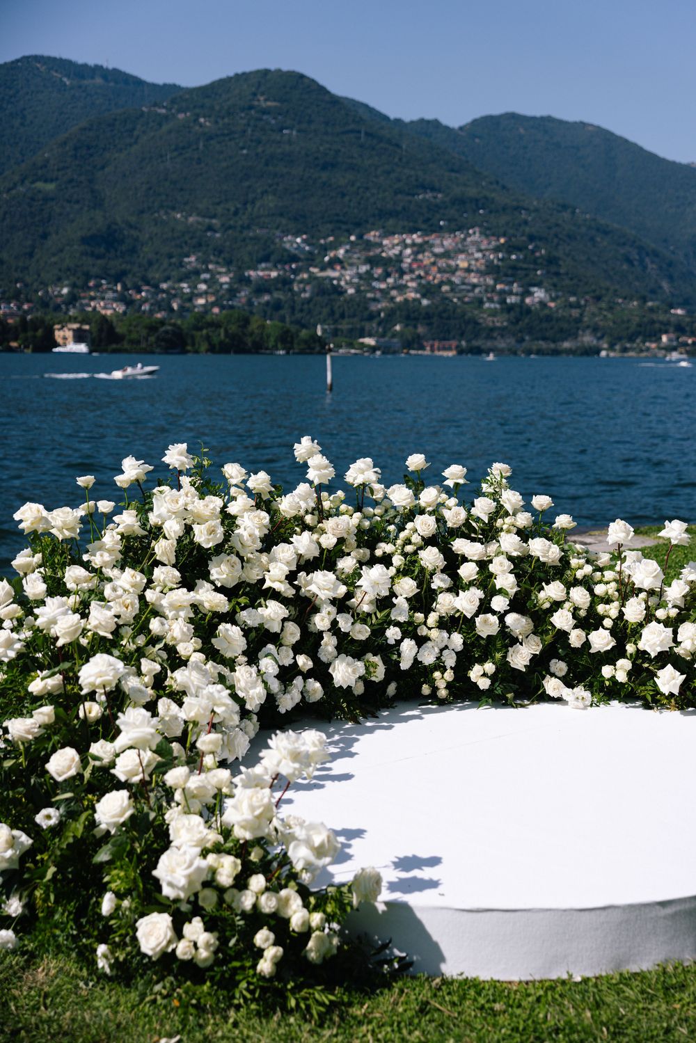 White rose arrangements line a lakeside ceremony setup with mountains and water in the background.