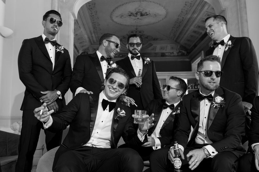 Group of groomsmen wearing sunglasses and tuxedos share drinks and laughs at a wedding celebration.