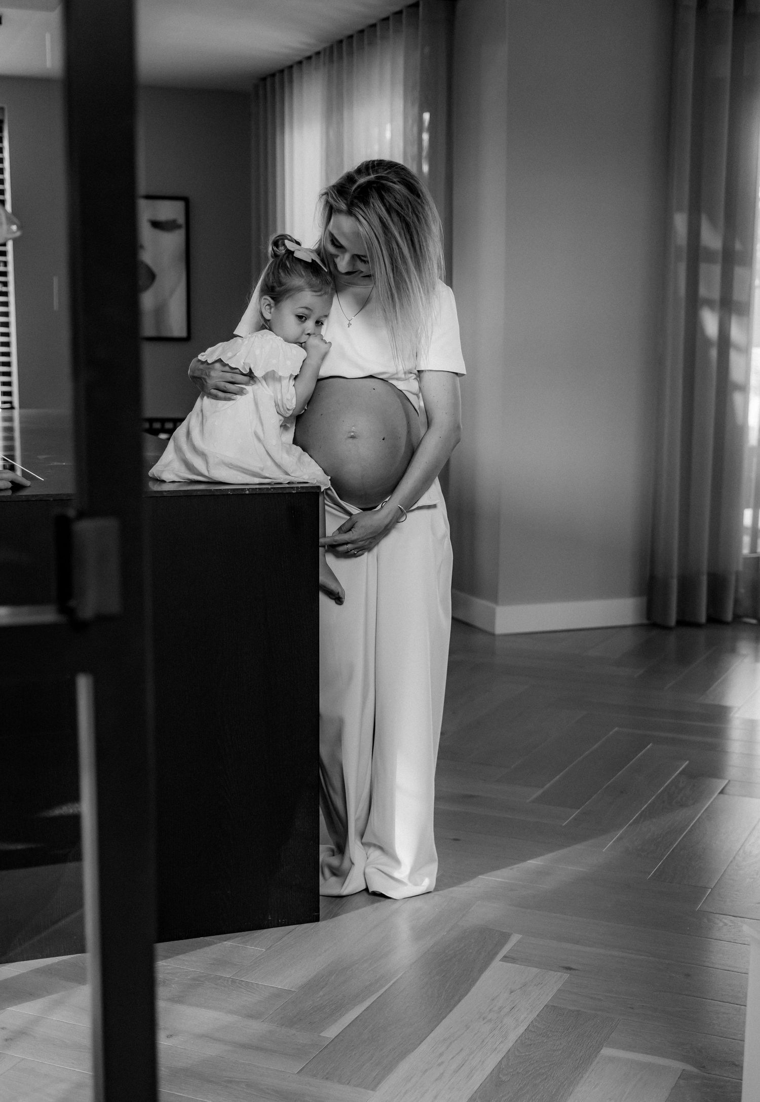 Black and white photo showing an expectant parent embracing a small child by a counter.