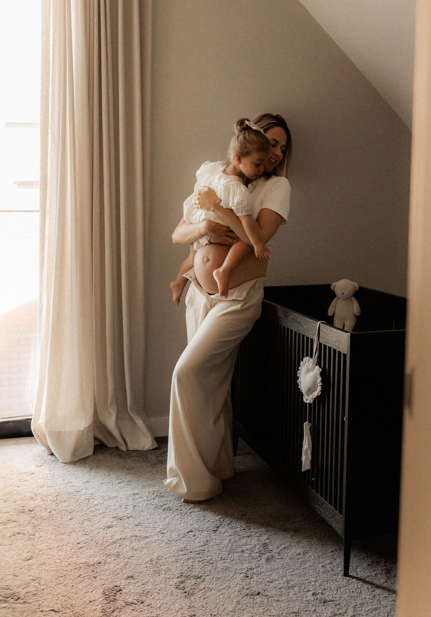 Parent holding child near a dark crib by window with flowing white curtains.