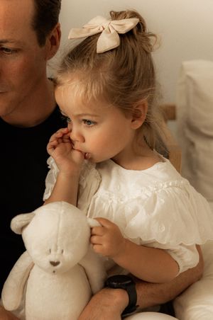 Small child in white dress holds stuffed bear while sitting with bow in hair.