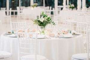 Elegant white wedding reception table setting with floral centerpiece and chiavari chairs in a bright venue.