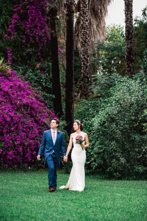 A couple walks together on a lush green lawn with vibrant magenta bougainvillea and palm trees in the background.