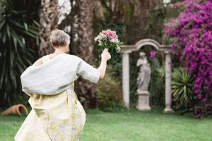 A person in a cream-colored dress holds a bouquet of pink roses in a garden with blooming bougainvillea and statues.