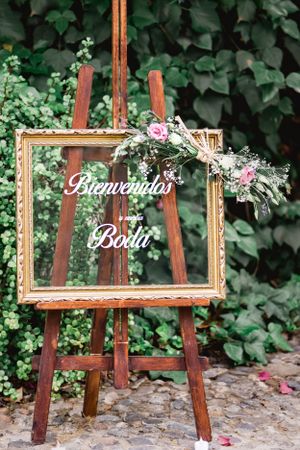 A wooden easel displays an ornate gold frame decorated with floral arrangements and 'Bienvenidos Bodas' text in a garden setting.