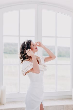 Elegant figure in white dress poses gracefully by bright arched windows in soft natural light.