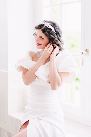 A bride in a white wedding dress and crystal headpiece smiles while touching her hair near a bright window.