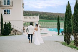 A couple walks away from a luxury villa with cypress trees and a swimming pool overlooking vineyard countryside.