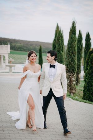 A couple walks together at a scenic venue with cypress trees, wearing formal wedding attire.