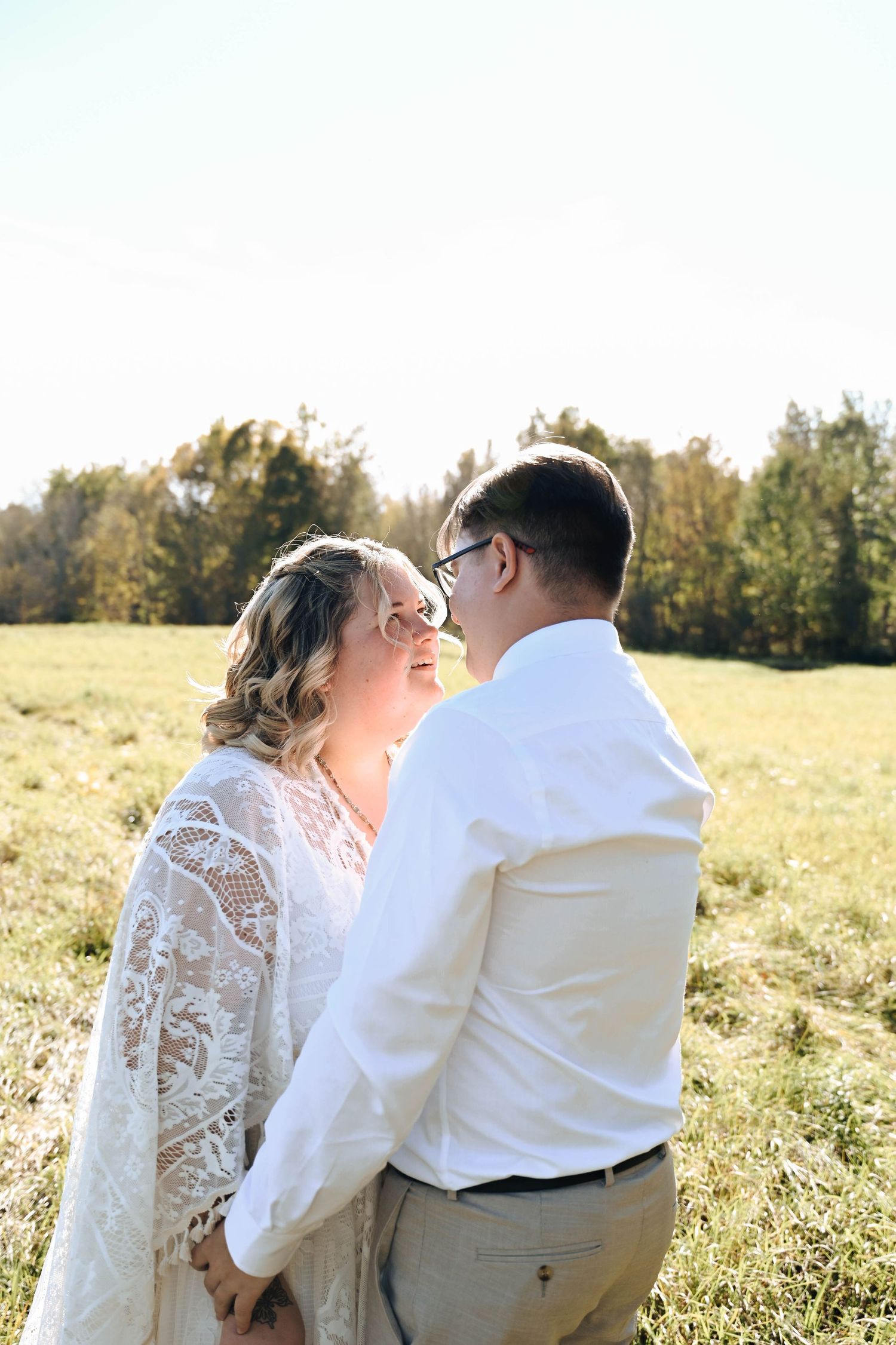 A happy couple shares a romantic moment in a sunlit meadow, with the bride wearing a lace dress and groom in white.