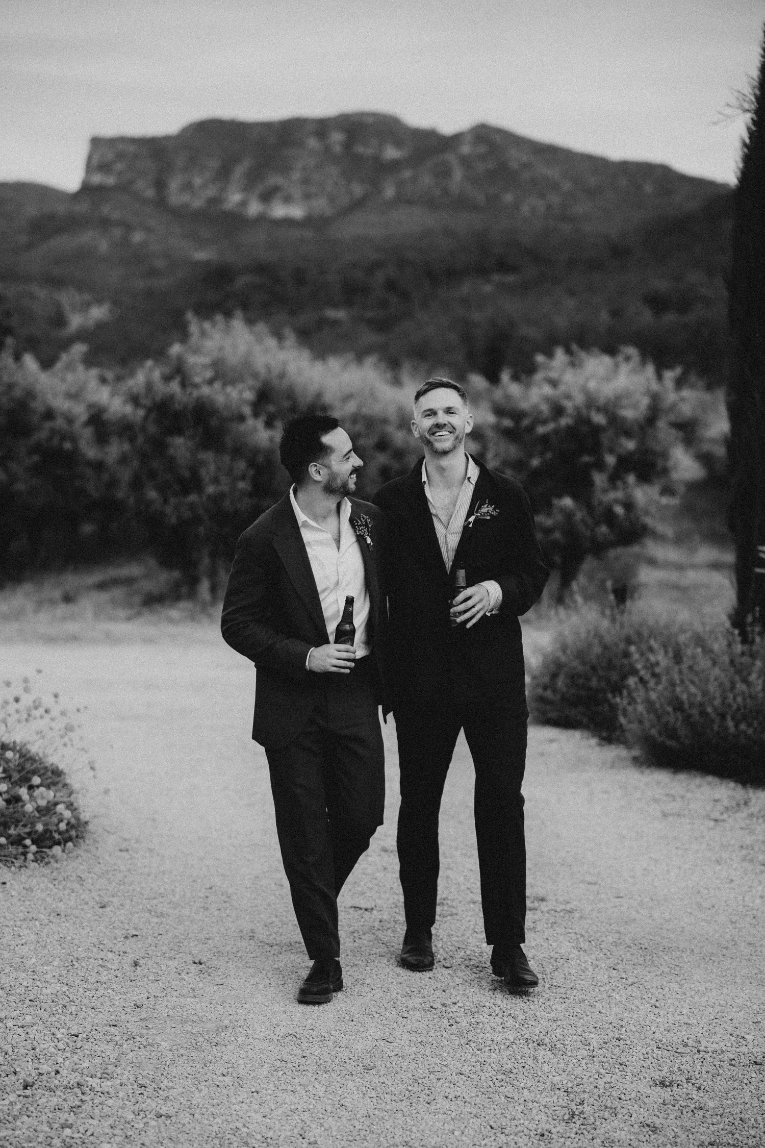 Two people in black suits share candid moments while walking along a desert path with mountains in the background.