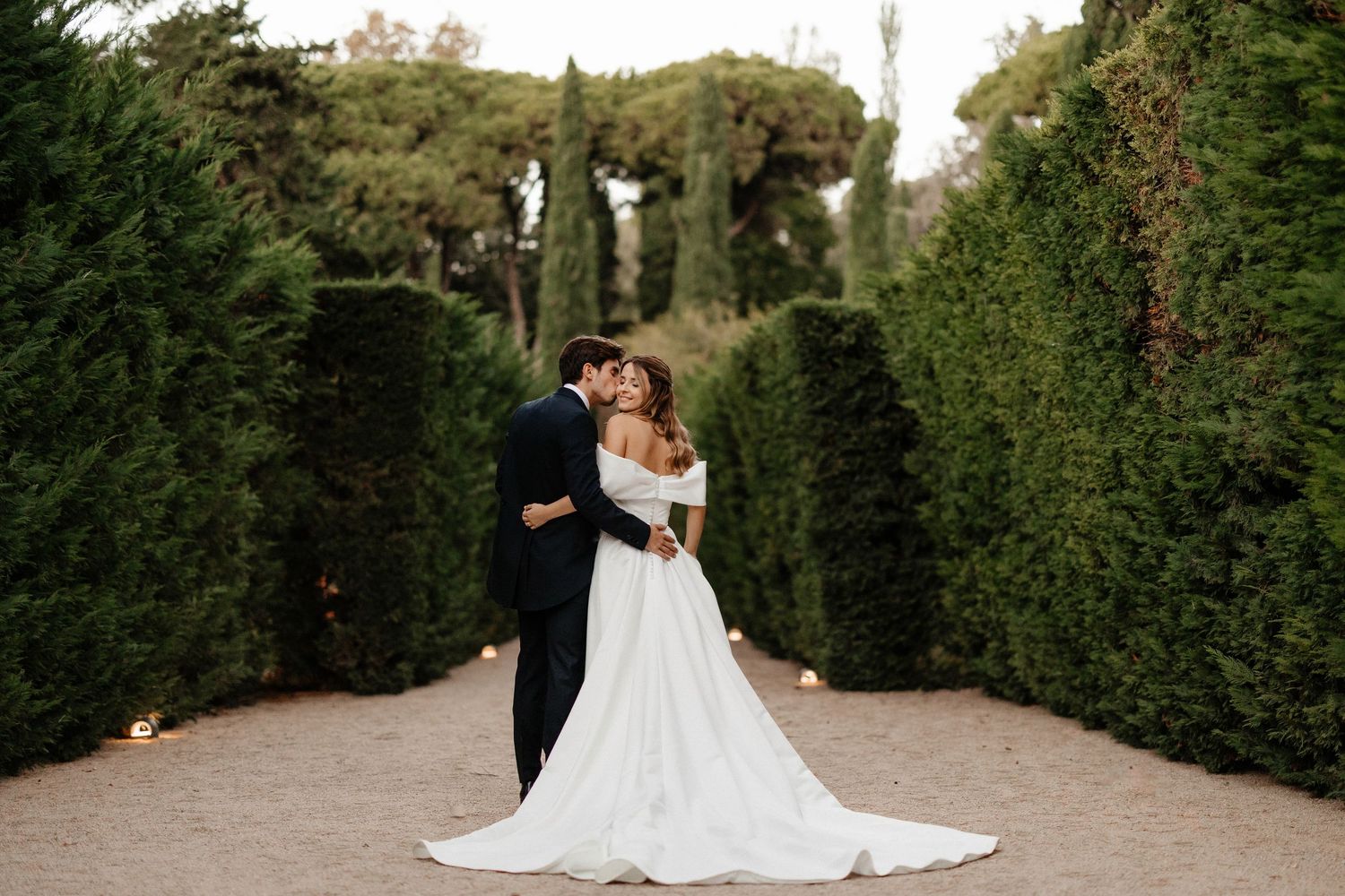 A couple embraces on a garden path surrounded by tall manicured hedges during their wedding photoshoot.