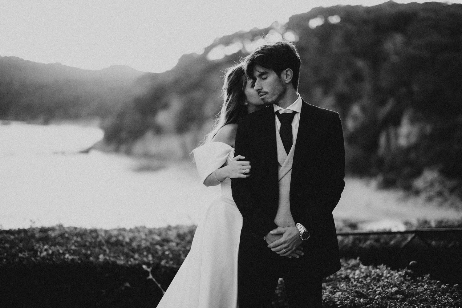 A romantic black and white wedding photo shows a couple embracing on a cliff overlooking a misty coastline.