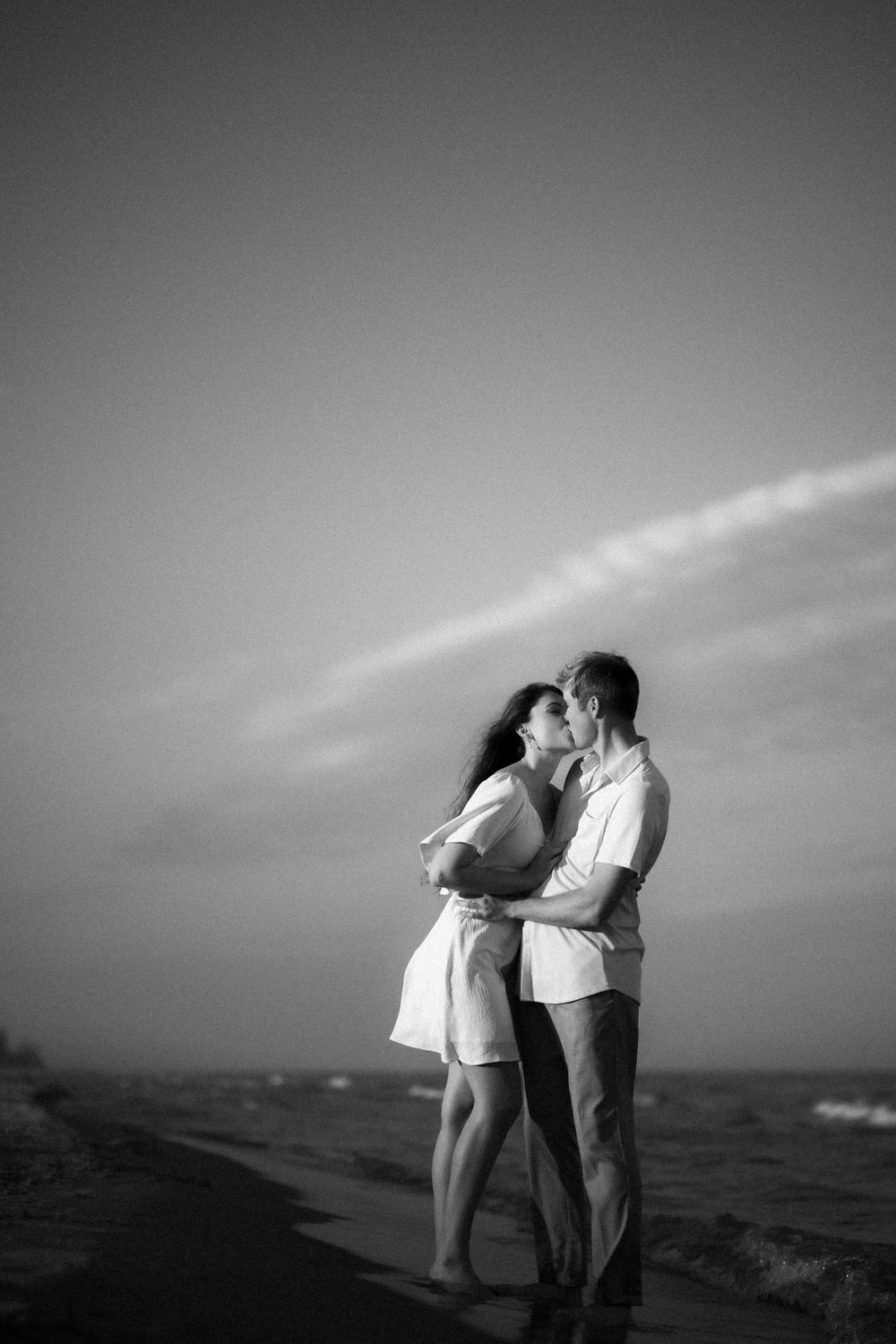 Black and white photo of couple in light summer clothing embracing on beach against dramatic sky.