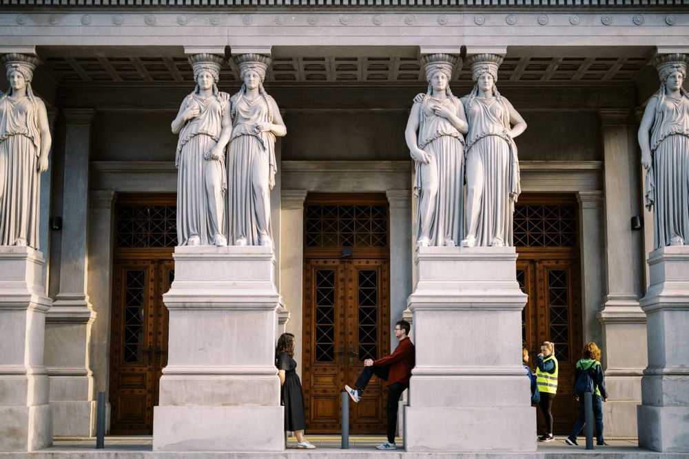 Ornate marble statues of robed figures stand guard at the entrance of a grand classical building with wooden doors.