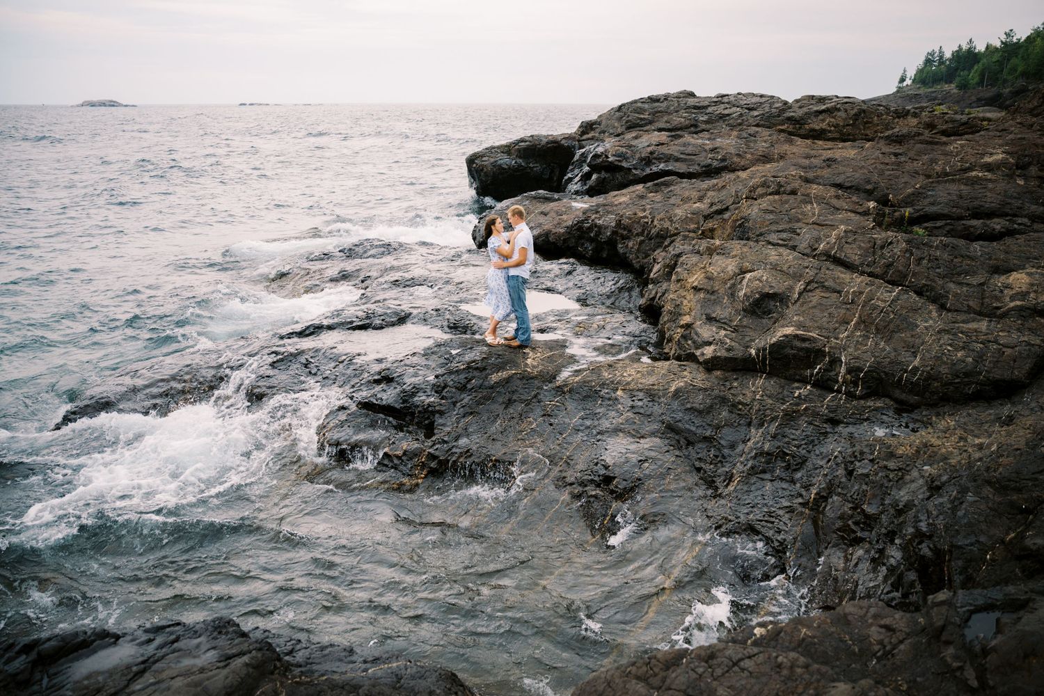 A couple embraces on rugged coastal rocks with crashing waves and ocean views.