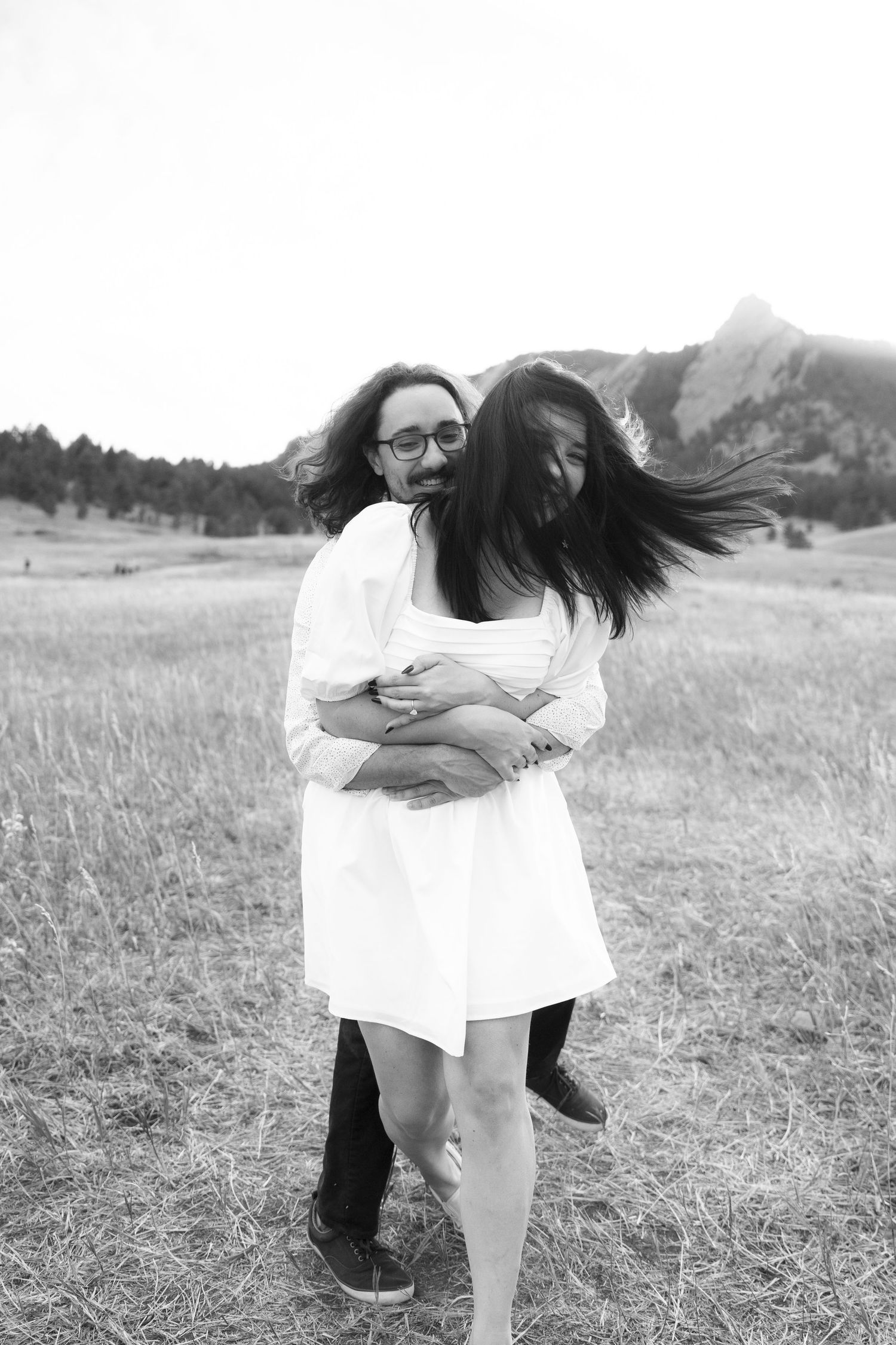 A playful black and white photo of a couple embracing in a field with mountains in the background.